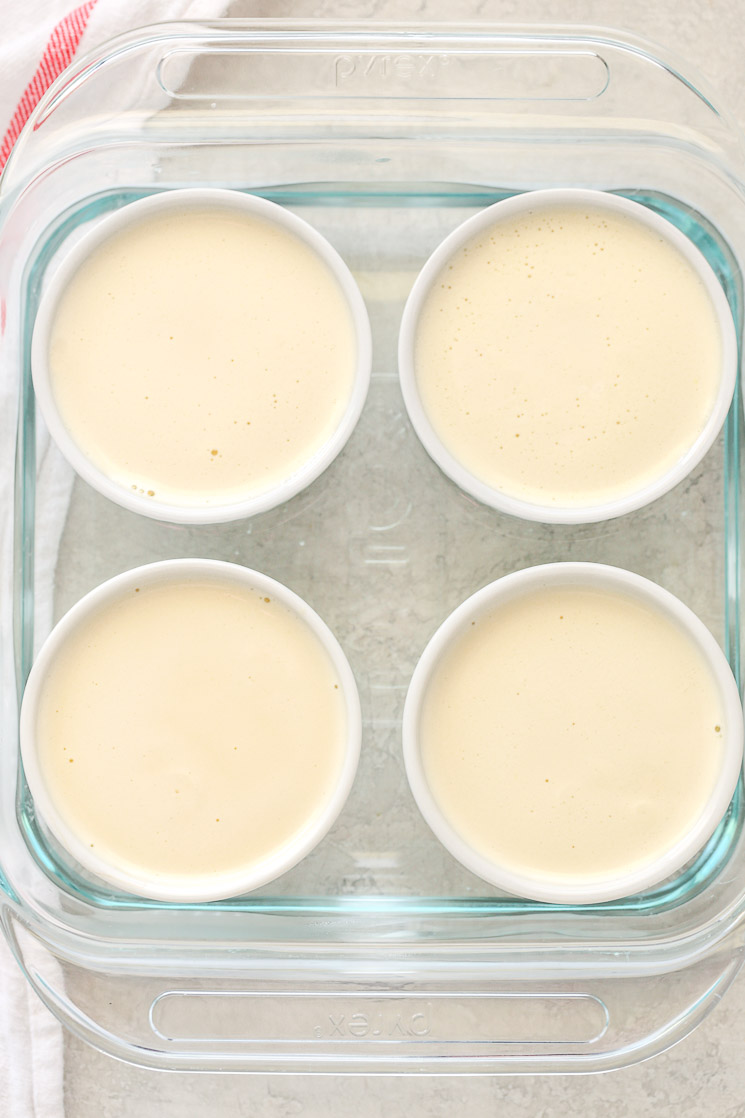 An 8-inch square glass baking dish with four white ramekins filled with the crème brûlée and water inside the dish.