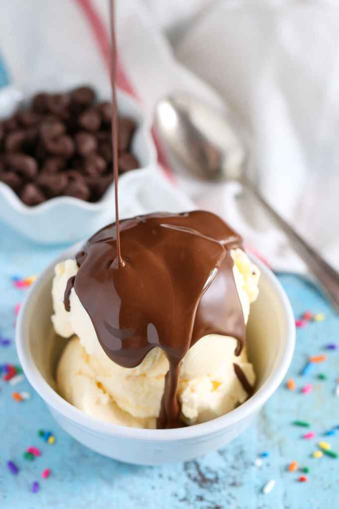 Magic shell ice cream topping being drizzled over a small dish of vanilla ice cream. A dish of chocolate chips and a spoon rest in the background. 