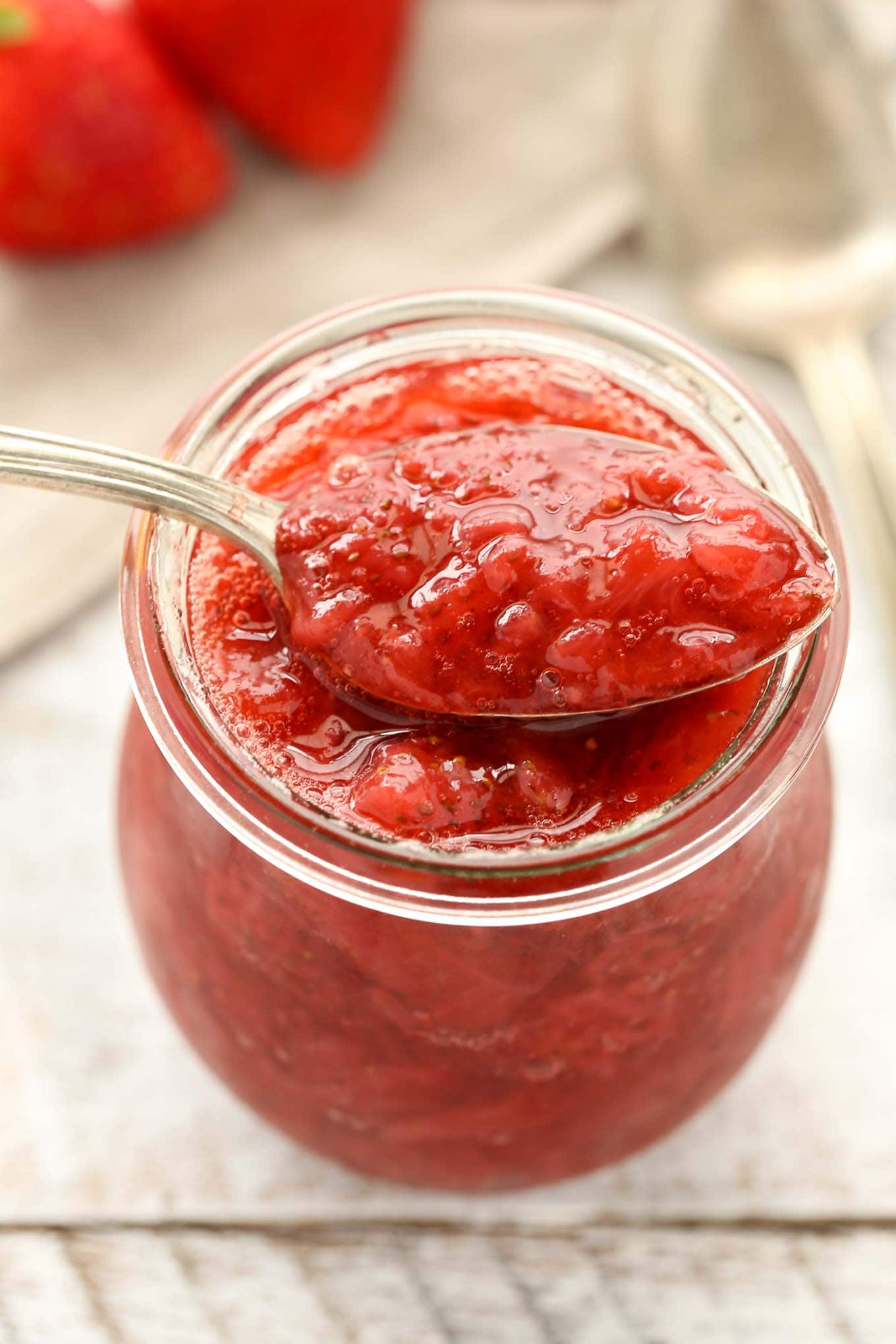 A spoon balanced on top of a glass jar of strawberry sauce. 