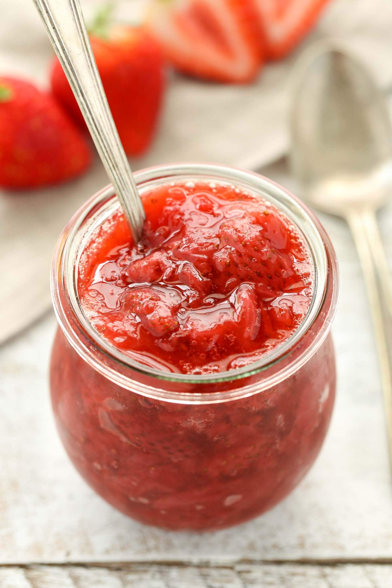 A glass jar full of strawberry topping that has a spoon handle sticking out of it. 