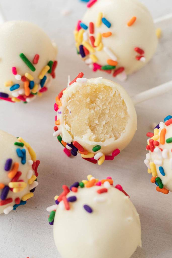 A close up view of vanilla cake pops topped with sprinkles. One cake pop has a bite missing. 