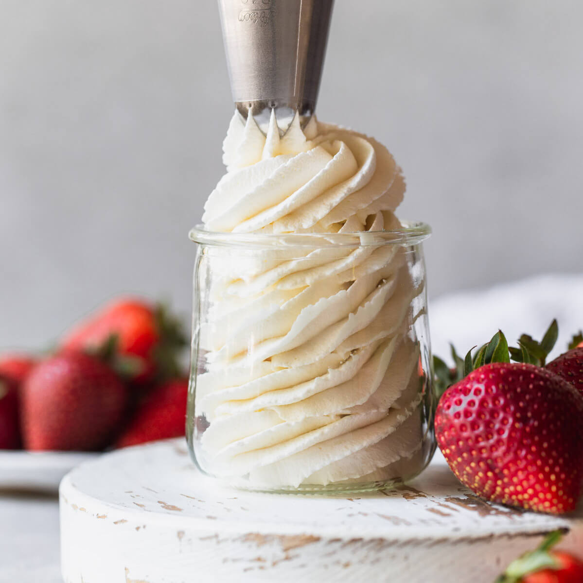 Homemade whipped cream being piped into a small glass jar surrounded by strawberries.