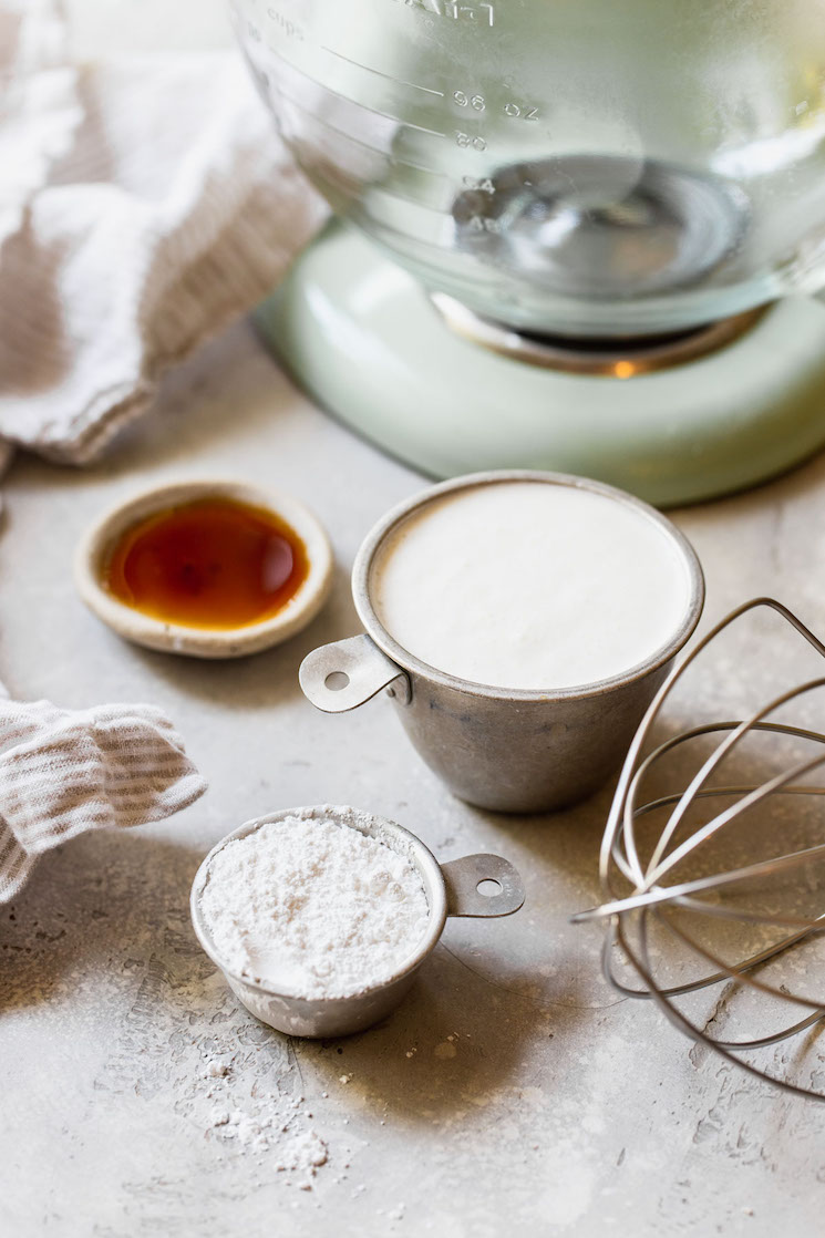 The ingredients for whipped cream sitting on top of a gray surface with a stand mixer in the background.