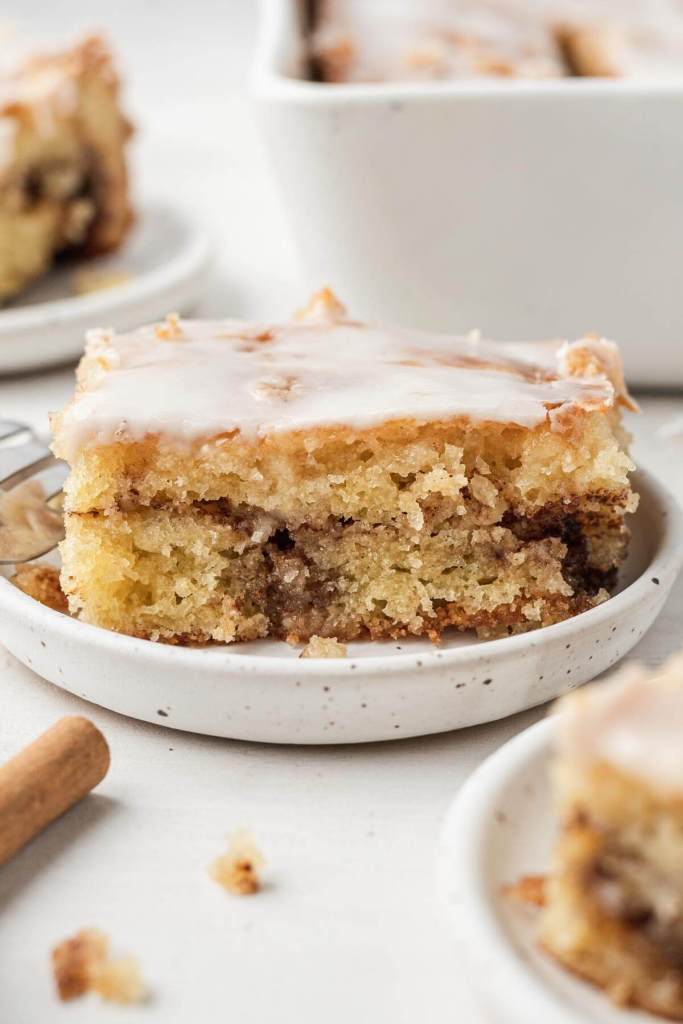 A slice of iced honey bun cake on a speckled white dessert dish. Additional cake slices rest in the foreground and background. 