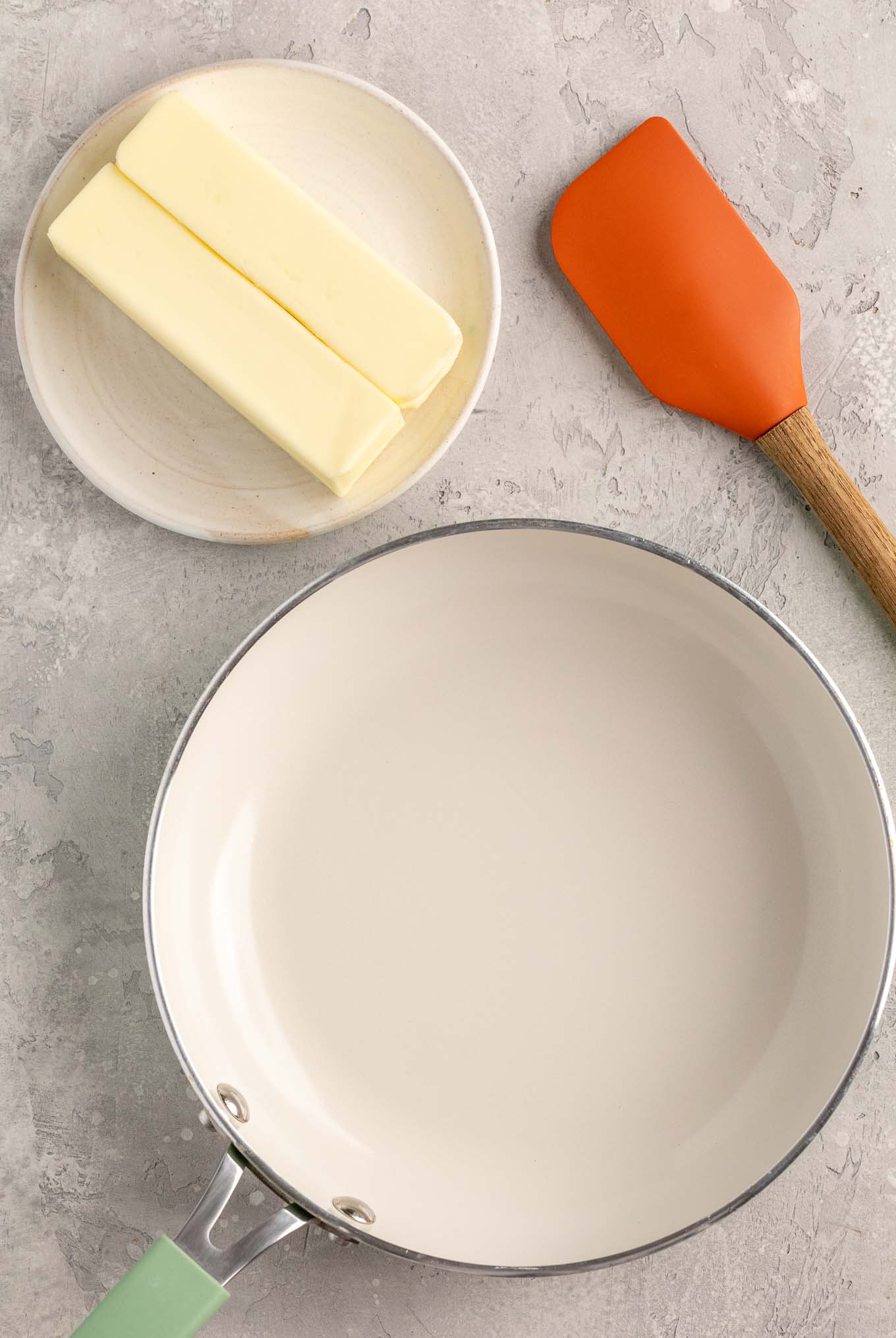 An overhead view of a white skillet, rubber spatula, and two sticks of butter on a white plate.