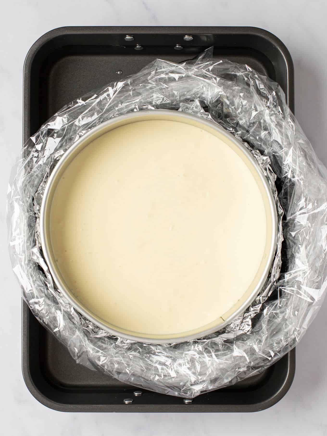 A cheesecake in a springform pan that's wrapped in foil and an oven bag. The wrapped pan is sitting inside of a large roasting pan with hot water.