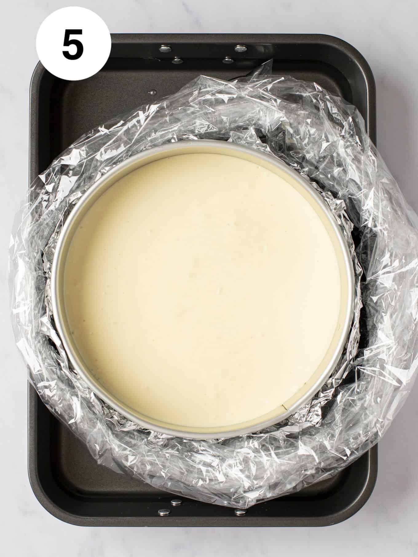 A cheesecake in a springform pan that's wrapped in foil and an oven bag and is sitting inside of a water bath.