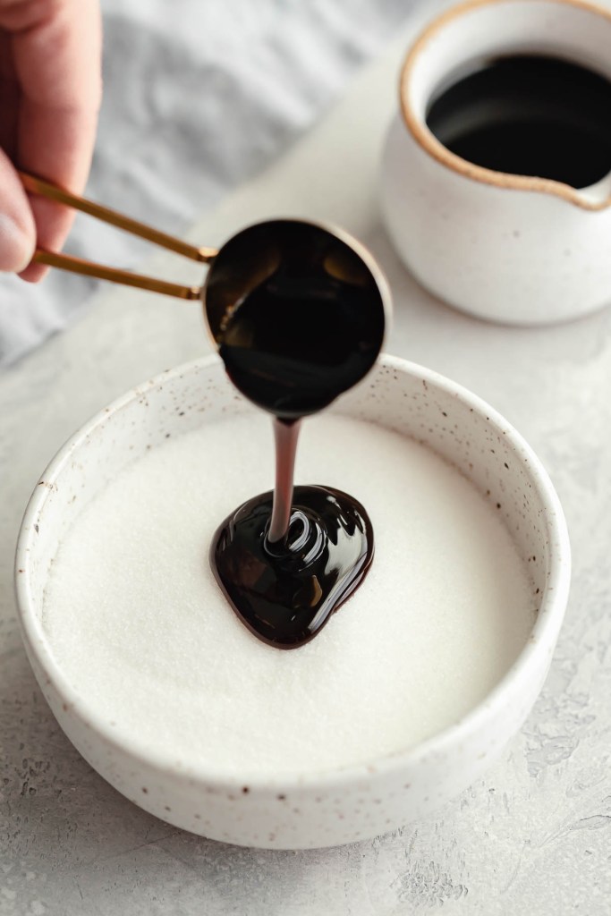 Molasses being poured into a bowl of granulated sugar. A small jug of molasses rests in the background. 