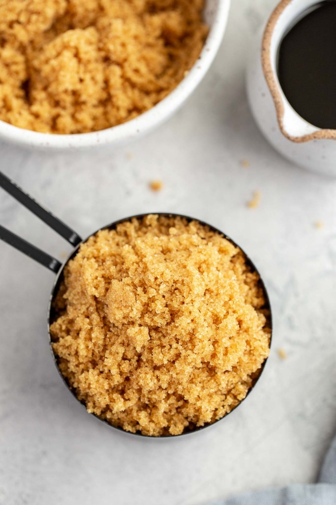 An overhead view of a measuring cup of homemade brown sugar. A bowl of brown sugar and a small jug of molasses rest nearby. 