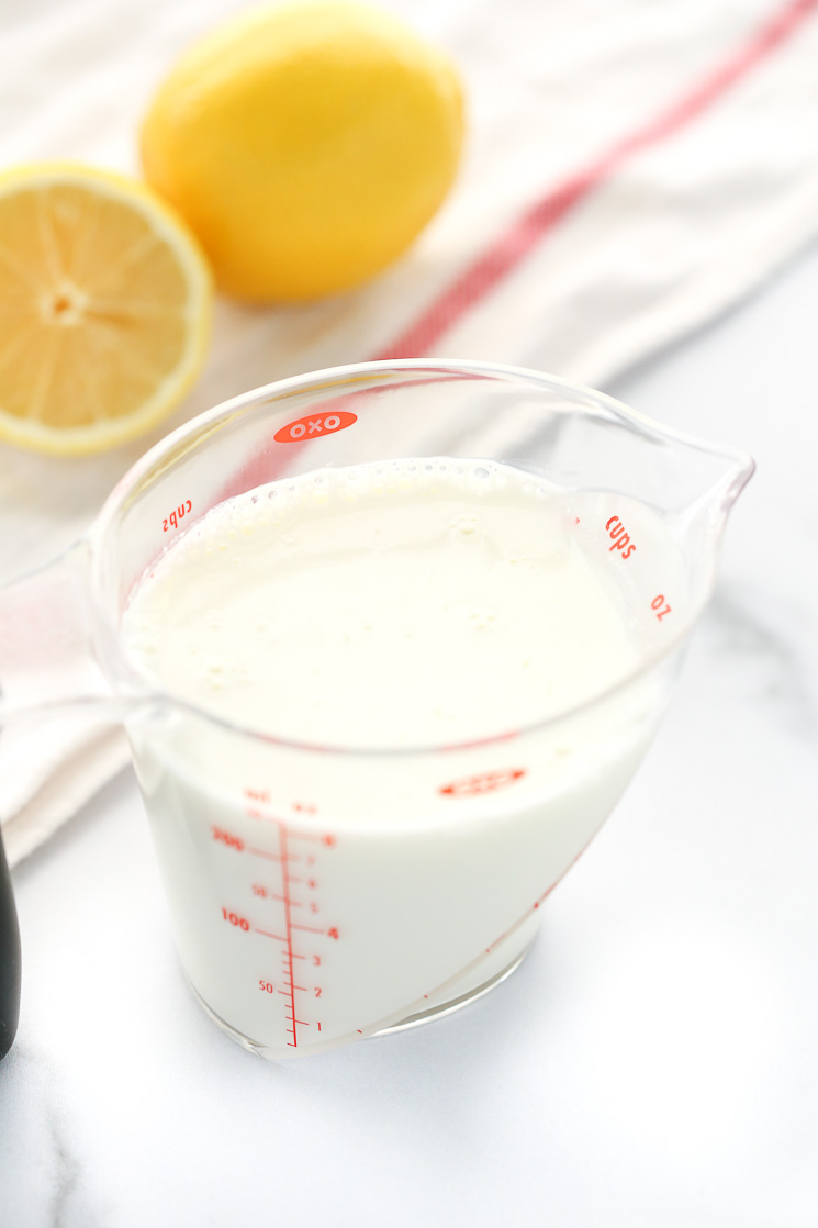 A clear measuring cup filled with homemade buttermilk along with a white napkin and lemons in the background.