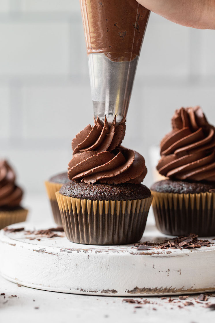 A rustic white board with chocolate cupcakes on it being topped with whipped chocolate ganache frosting.