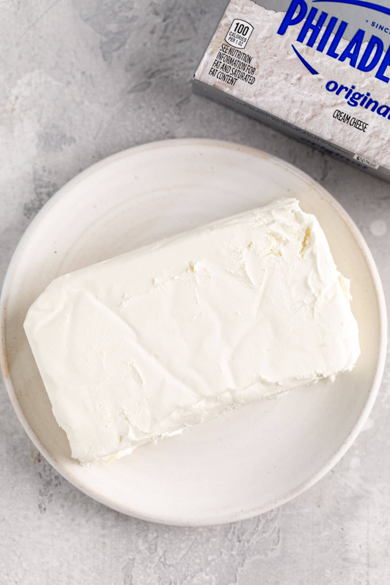 A block of cream cheese on a white plate. A box of Philadelphia cream cheese is sitting next to the plate.