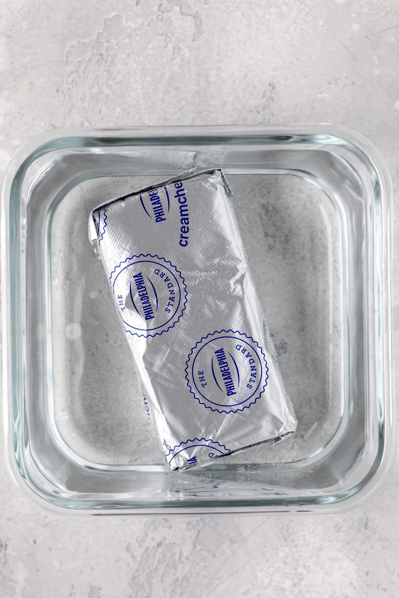 An overhead view of a sealed package of cream cheese in a small container filled with warm water.