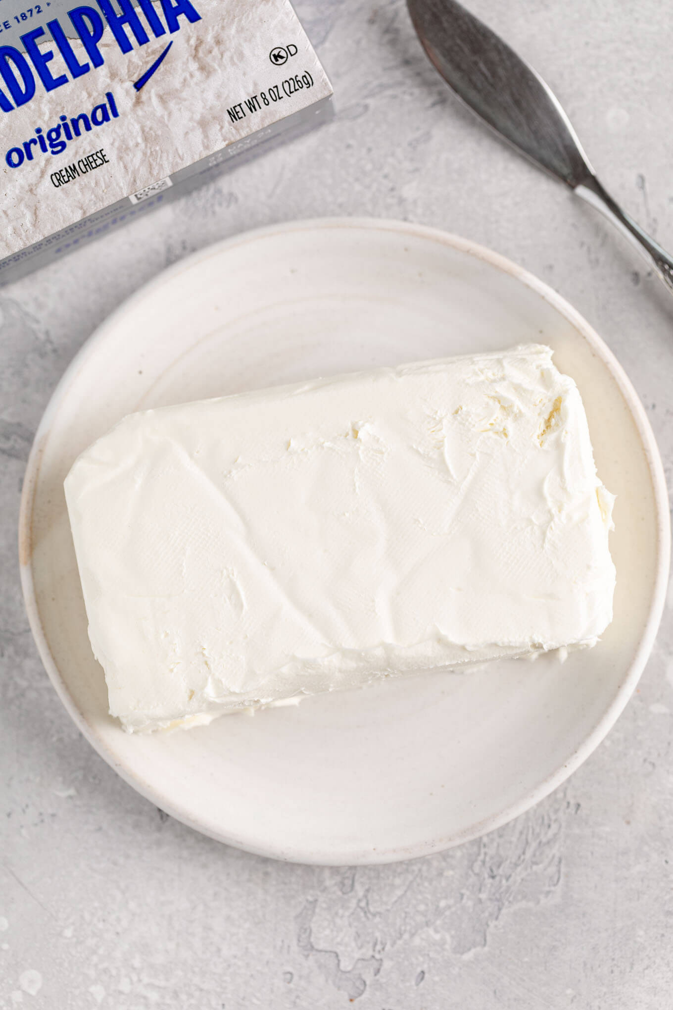 A block of cream cheese on a white plate. A box of Philadelphia cream cheese is sitting next to the plate.