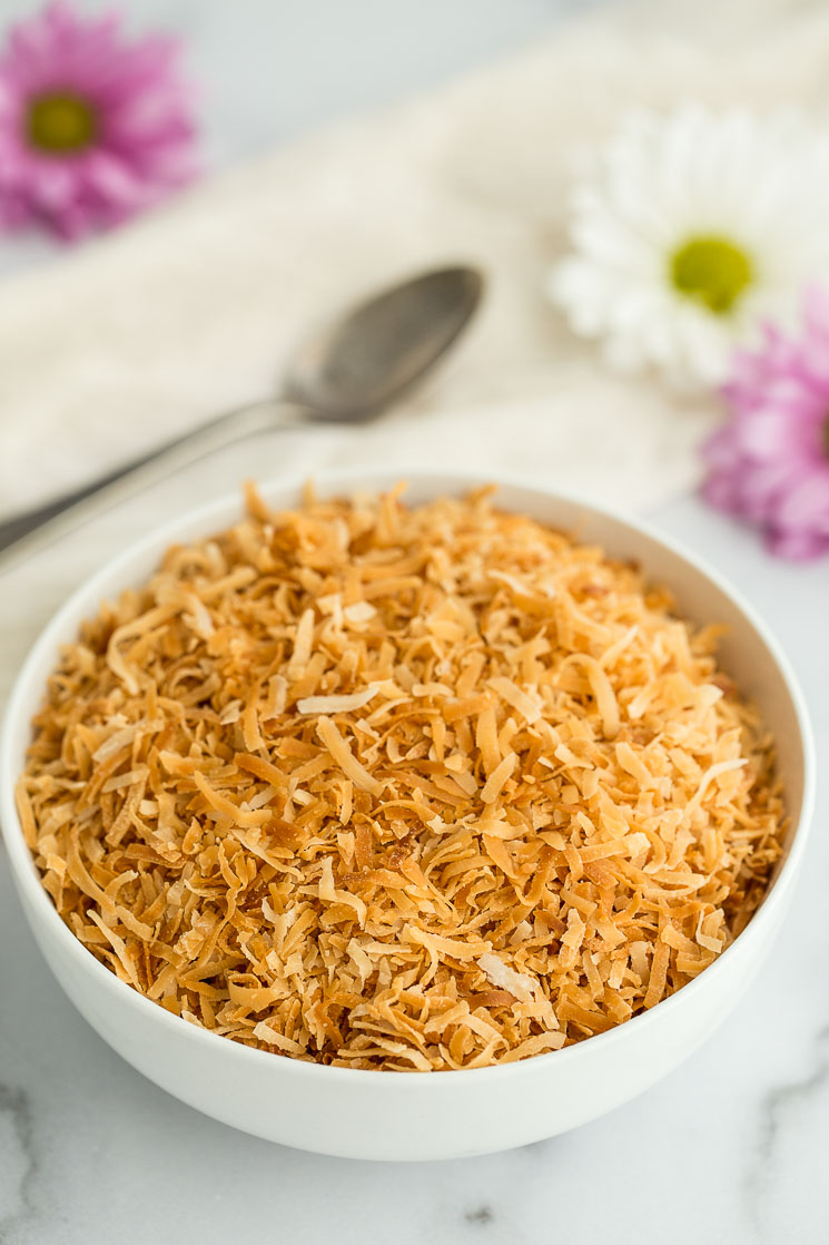 A white bowl filled with toasted coconut with an antique spoon and flowers in the background. 