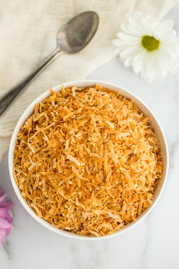 A white bowl filled with toasted coconut with an antique spoon and flowers in the background. 