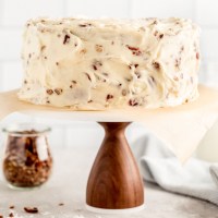 An Italian cream cake on a wooden cake stand with a marble top. A jar of chopped pecans and two milk jugs rest in the background.