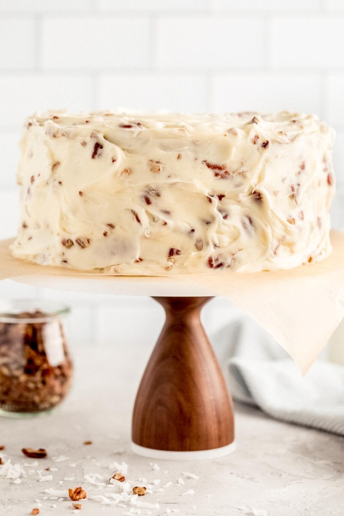 An Italian Cream Cake on top of a wooden cake stand with a marble top. A jar of chopped pecans and two milk jugs rest in the background.