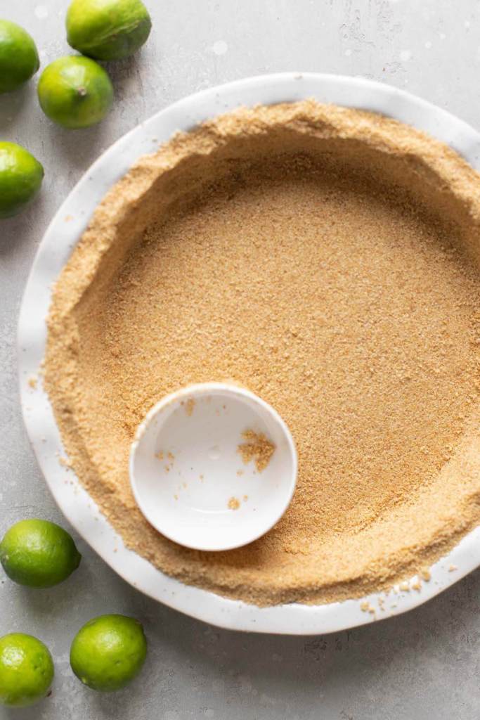 Overhead view of a graham cracker crust in a white pie plate, being packed down with a measuring cup. 