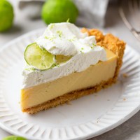 A slice of key lime pie on a white dessert plate. Key limes are scattered in the background.