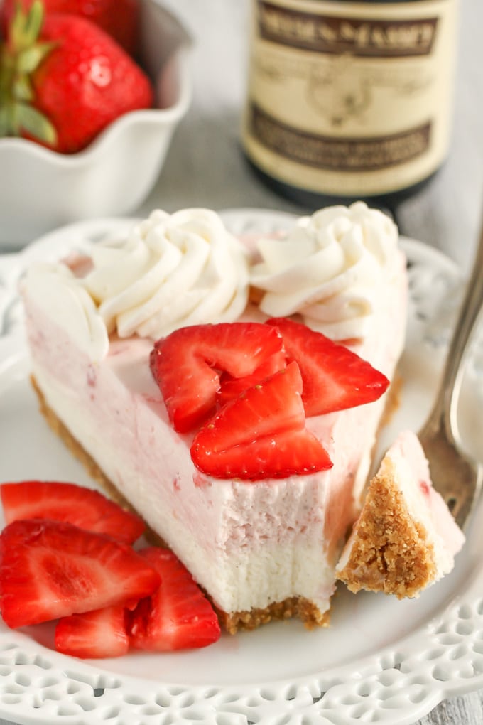 A slice of no bake strawberry cheesecake on a white dessert plate with berry slices and a fork. A bottle of vanilla extract rests in the background. 