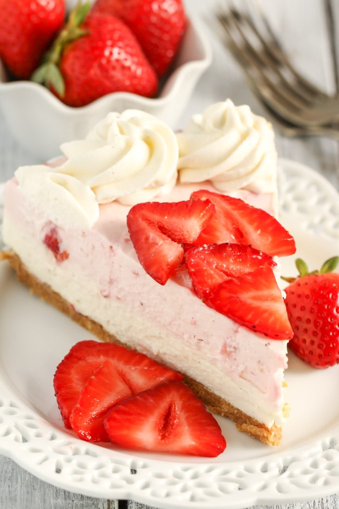 A slice of no bake strawberry cheesecake on a white dessert plate with berry slices. A dish of berries and a fork rest in the background. 