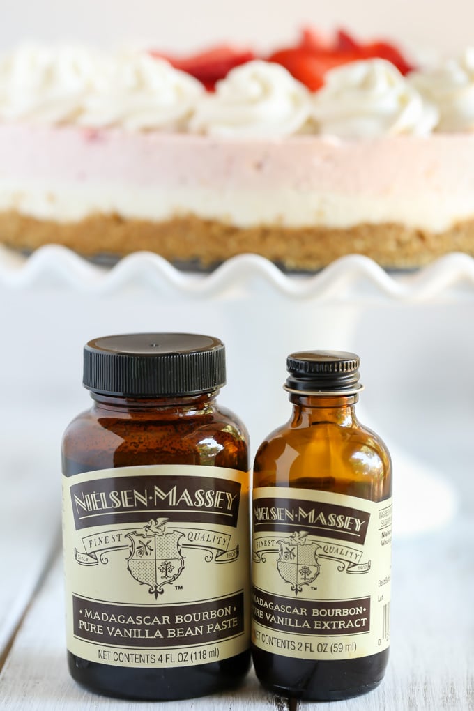 A bottle of vanilla bean paste and a bottle of vanilla extract. In the background is a fresh strawberry cheesecake on a cake stand. 