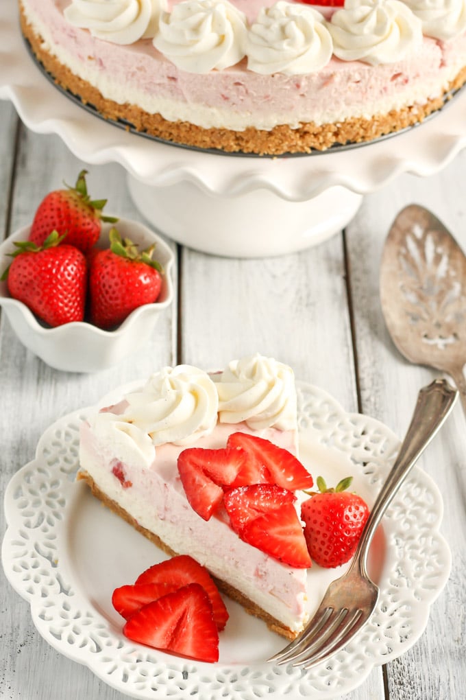 A slice of no bake strawberry cheesecake on a white dessert plate with a fork. A small dish of fresh strawberries and the remainder of the cheesecake rest in the background. 