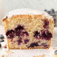 A straight on view of a loaf of lemon blueberry bread on a piece of parchment paper. The bread has been sliced to show the interior texture. A bowl of blueberries rests in the background.