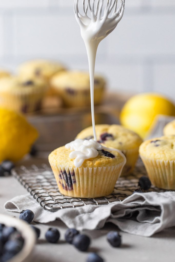 A whisk being held over a lemon blueberry muffin. Glaze drizzles down from the whisk. Additional muffins are in the background of the image and fresh blueberries are in the foreground. 