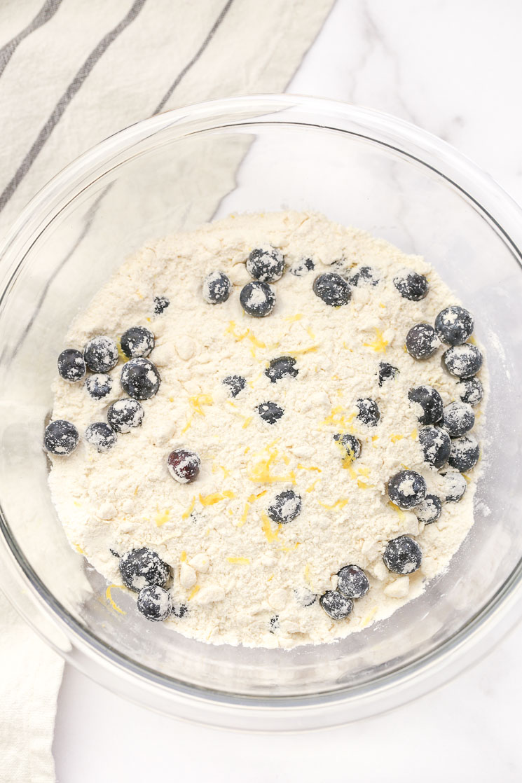 A glass bowl filled with the dry ingredients, blueberries, and lemon zest for scones and a light gray napkin on the side.