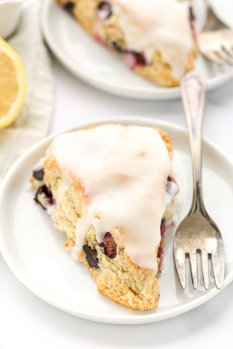 A blueberry scone with lemon glaze on a white plate with another scone and sliced lemon in the background.