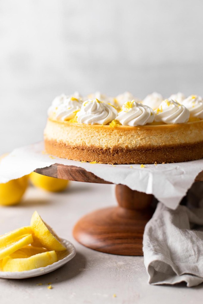 A side view of a lemon cheesecake on a wooden cake stand. Lemon slices rest on a small plate nearby. 