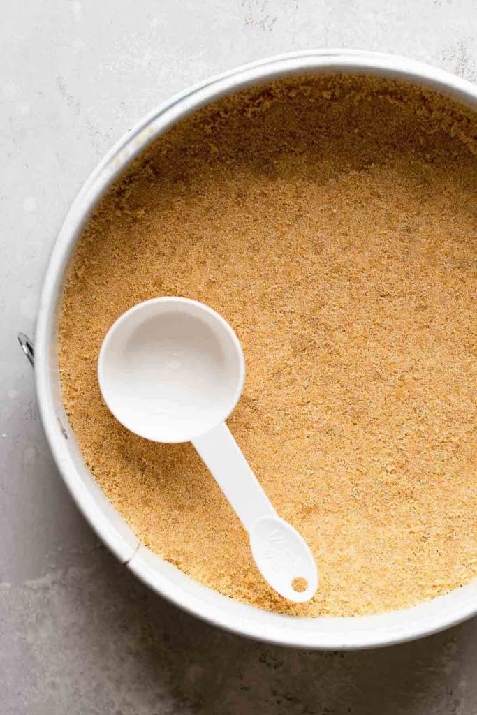 An overhead view of a graham cracker crust in a springform pan. A measuring cup rests on the crust. 