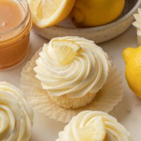 Lemon cupcakes topped with lemon buttercream and a lemon wedge. A bowl of lemons and jar of lemon curd are sitting next to the cupcakes.