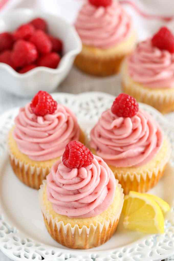 Lemon cupcakes topped with raspberry frosting and fresh raspberries on a white plate. A bowl of berries and more cupcakes rest in the background. 