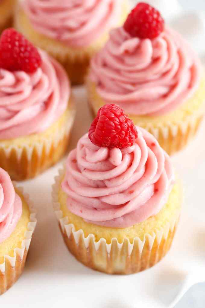 Close up of lemon cupcakes topped with raspberry buttercream and fresh raspberries on a white platter.