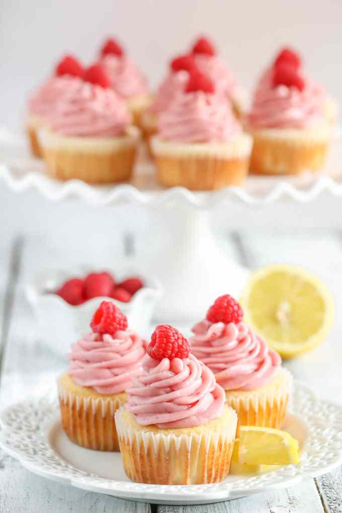 Lemon cupcakes topped with raspberry frosting and fresh raspberries on a white plate. More cupcakes rest on a cake stand in the background. 