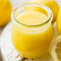 A small glass jar filled with lemon curd on top of a worn white wood surface.