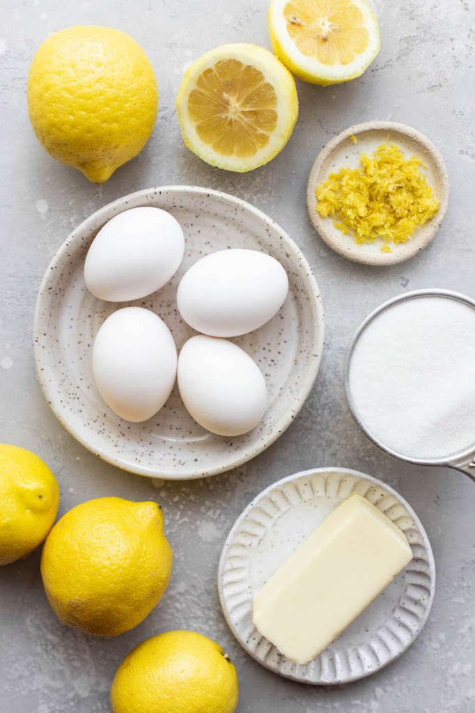 Eggs, lemons, lemon zest, butter, and sugar in various bowls and plates on a gray surface.