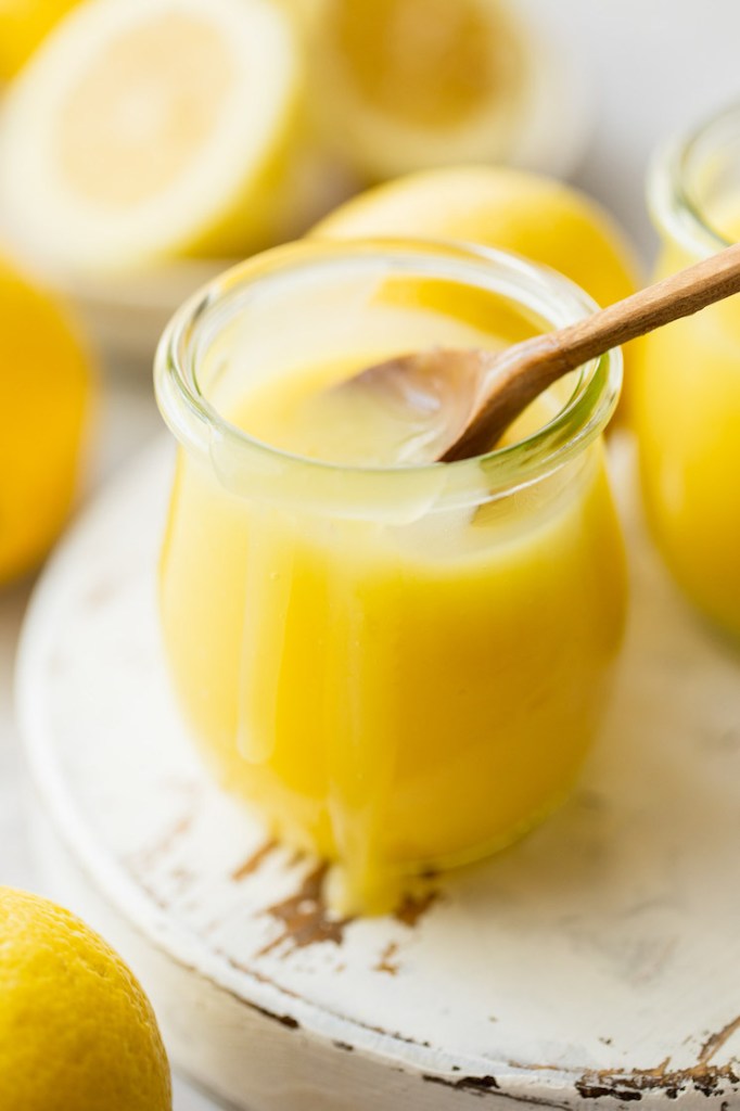 A wooden spoon resting in a glass jar filled with lemon curd on a rustic wood surface.