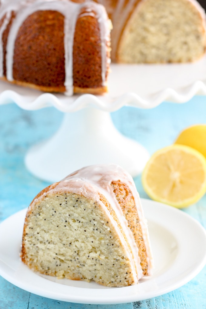 A slice of glazed lemon poppy seed bundt cake on a white plate. The rest of the cake is on a cake stand in the background. 