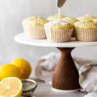 A marble cake stand with lemon poppy seed muffins being topped with a lemon glaze.