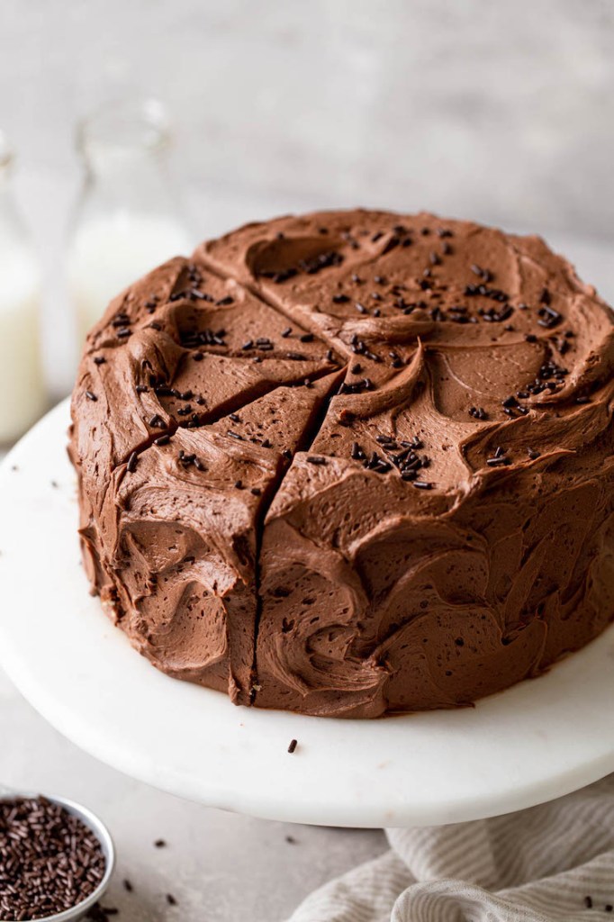 A chocolate marble cake on a white cake stand, topped with chocolate sprinkles. Two glass milk jugs rest in the background. 