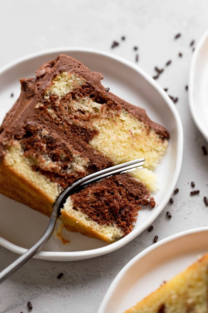 An overhead view of a slice of marble cake on a white plate. A fork is digging into the slice. 