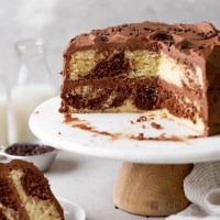 A side view of a homemade marble cake on a cake stand. A slice has been removed and has been plated. Milk jugs rest in the background.