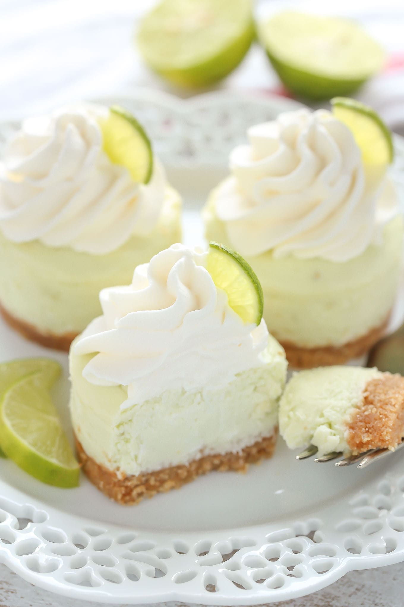 A white plate with three mini key lime cheesecakes on it. A lime wedge rests on one side and a fork rests on the other. 