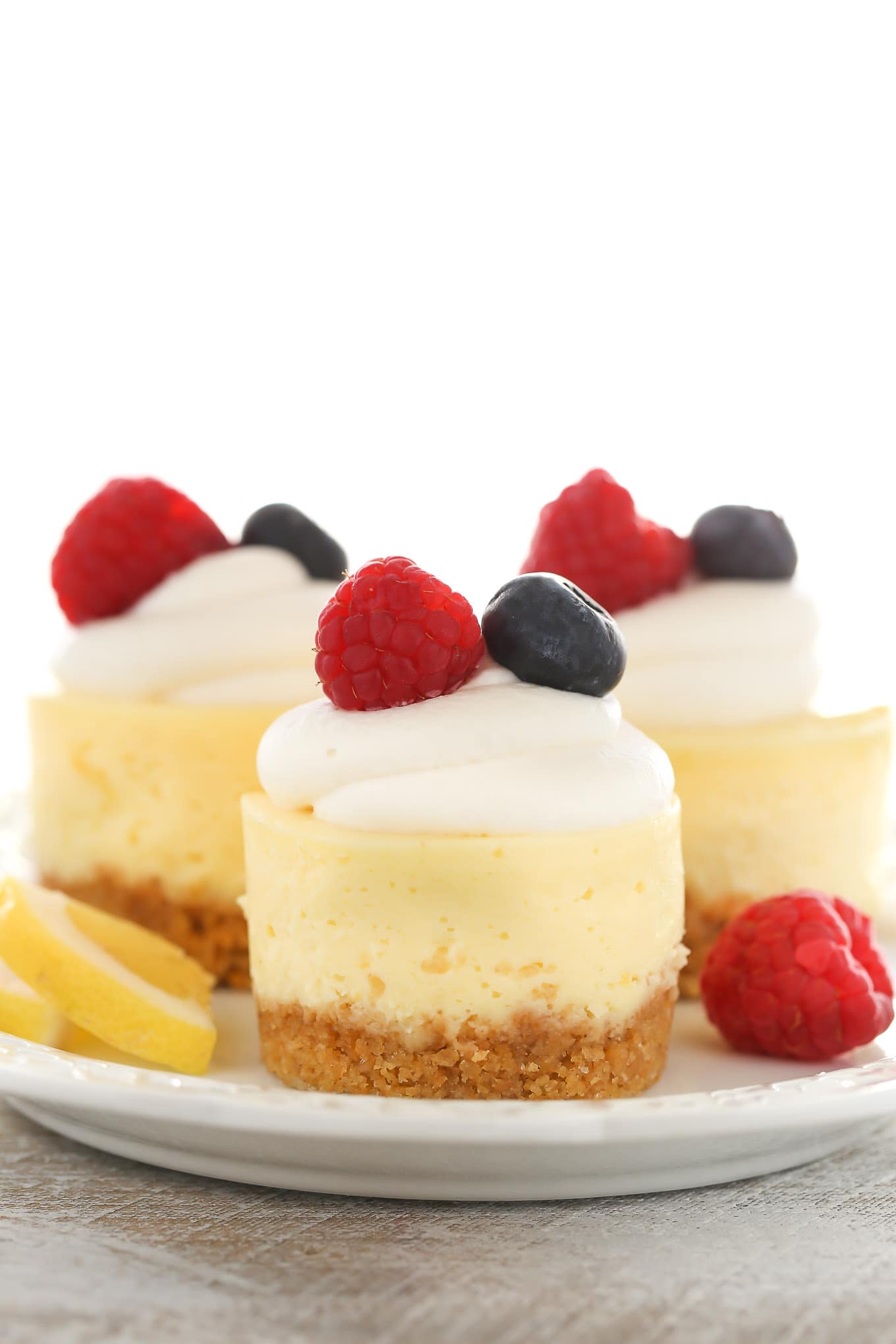 A side view of three lemon cheesecake bites on a white plate. Each is topped with whipped cream and fresh berries. 