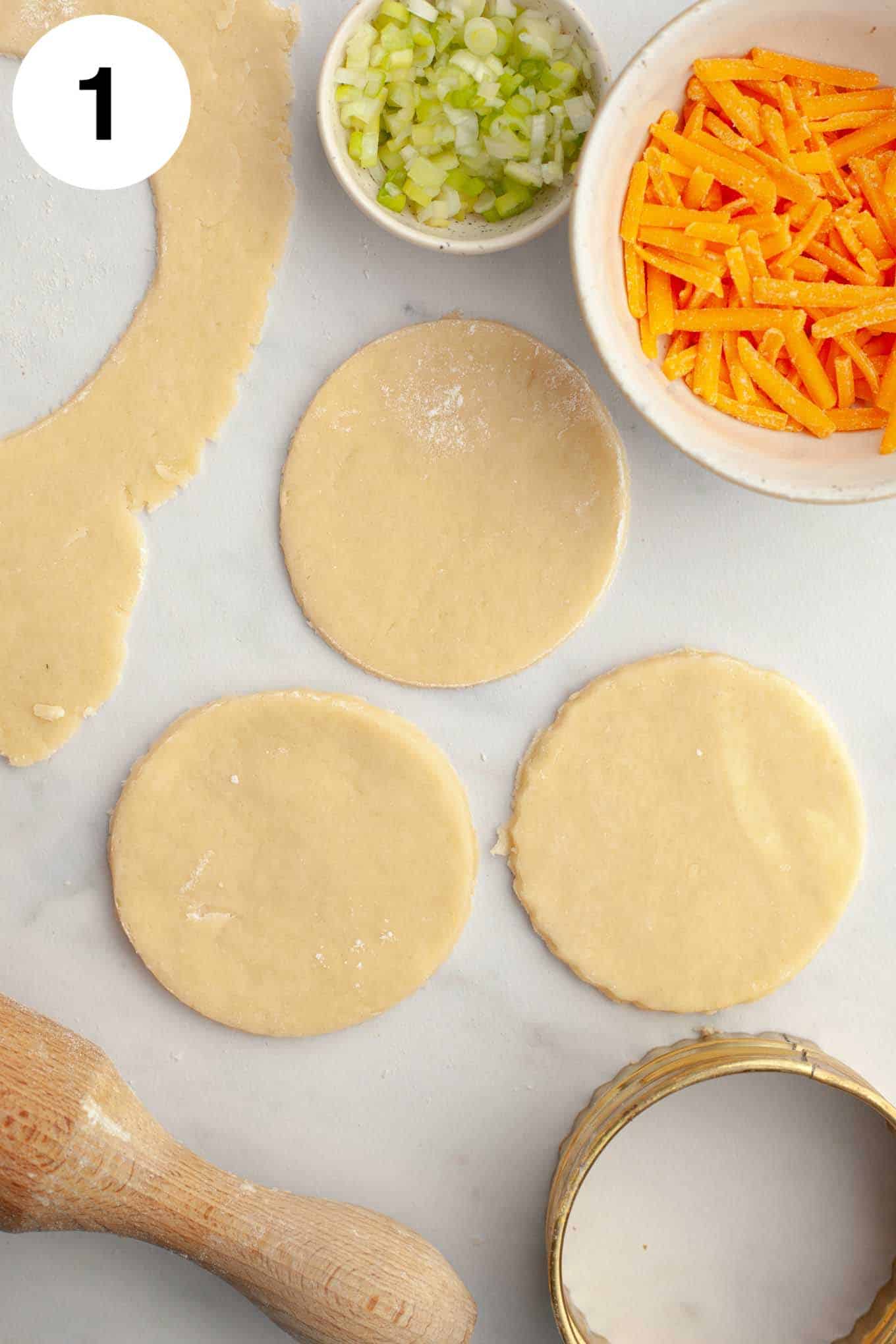 An overhead view of three mini pie crust circles with quiche filling ingredients portioned into bowls. 