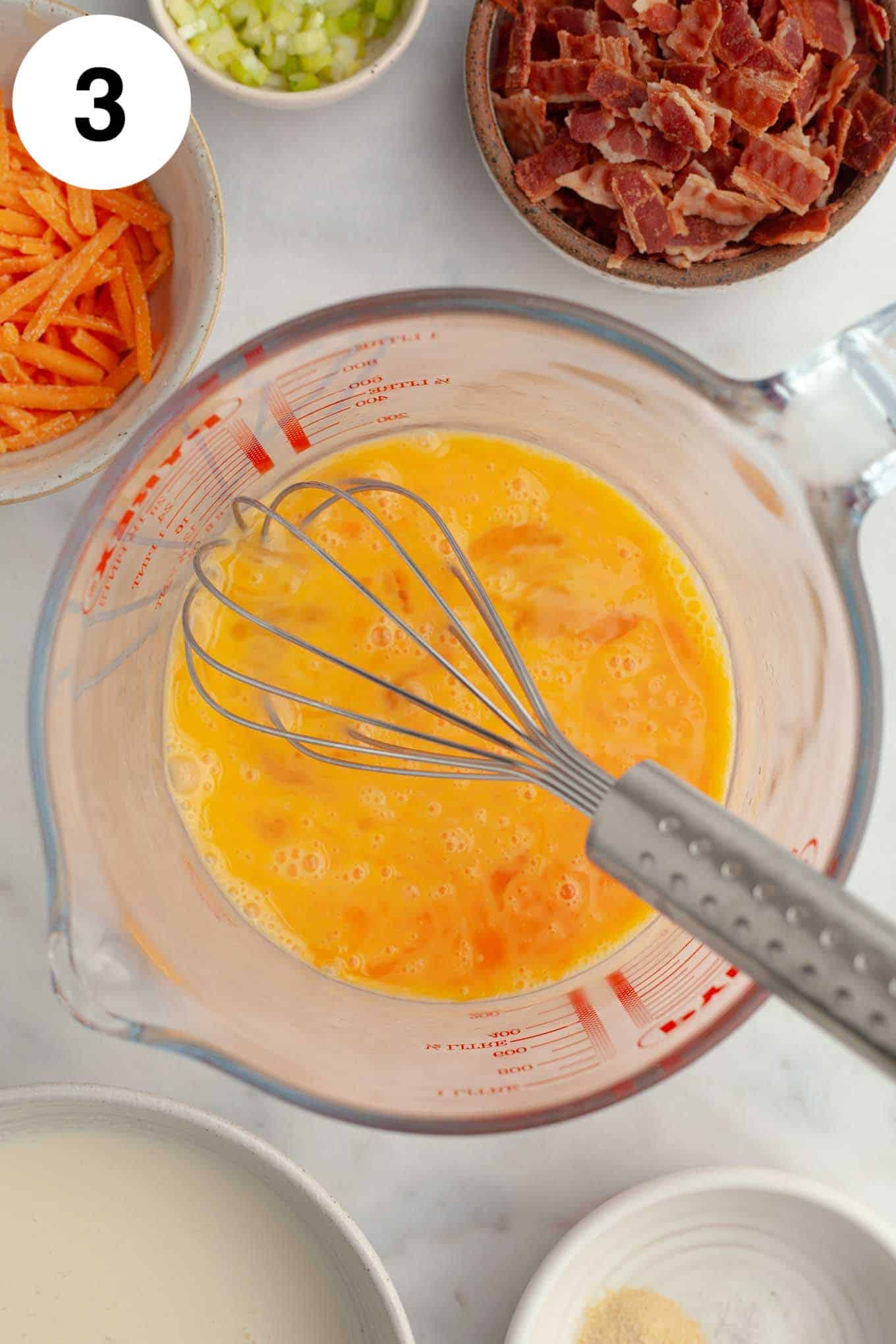 An overhead view of beaten eggs in a measuring cup, with bowls of quiche mix-ins nearby. 